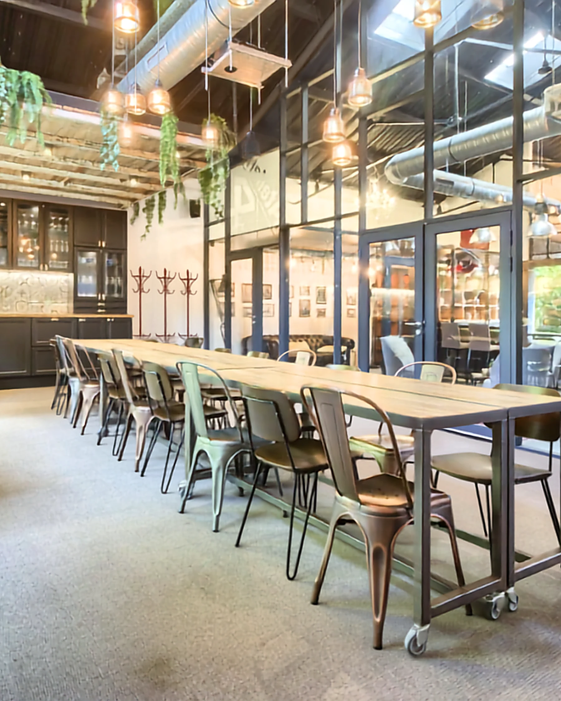 Intérieur d'un espace de coworking de style industriel avec une longue table en bois, des chaises en métal coloré et des luminaires suspendus, paroi vitrée noire séparant les espaces